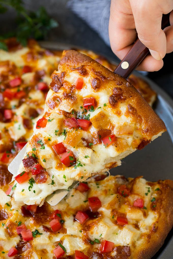 Una mano sosteniendo una espátula con un trozo de pizza Chicken Alfredo.