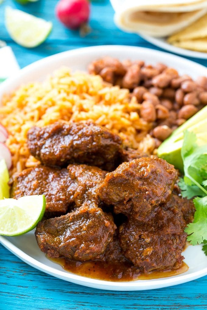 Esta receta de chile de Colorado es un platillo tradicional mexicano hecho con tierna carne de res cocinada a fuego lento en una sabrosa salsa de tomate.  ¡Es el acompañamiento perfecto para burritos, enchiladas y tacos! Chile Colorado en un plato servido con arroz y frijoles.