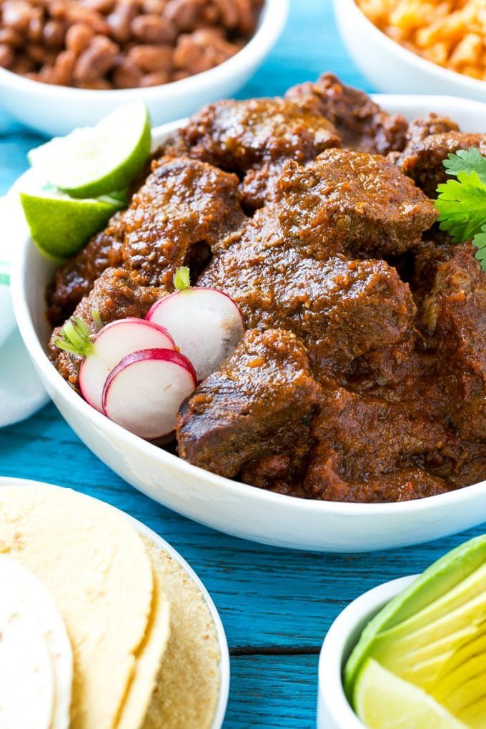 Esta receta de chile de Colorado es un platillo tradicional mexicano hecho con tierna carne de res cocinada a fuego lento en una sabrosa salsa de tomate.  ¡Es el acompañamiento perfecto para burritos, enchiladas y tacos! Un tazón de chile colorado servido con tortillas, frijoles y aguacate.