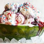 Esta receta de helado de arándano blanco es una sandía cargada con tres tipos de helado, salsas caseras de fresa y arándano, una cobertura de malvavisco, y todo termina con crema batida, chispas y chispas de fresa cubiertas de chocolate.  ¡Es el espectáculo perfecto del 4 de julio!