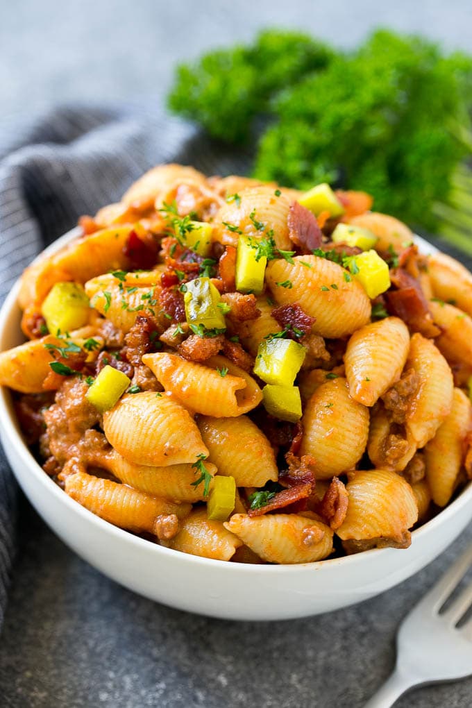 Un tazón de pasta de hamburguesa con queso cubierto con tocino picado y encurtidos.