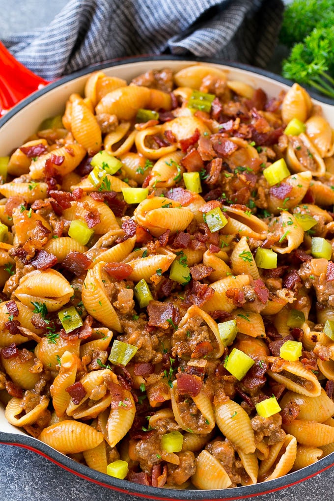 Pasta de hamburguesa con queso en una sartén adornada con pepinillos y tocino.