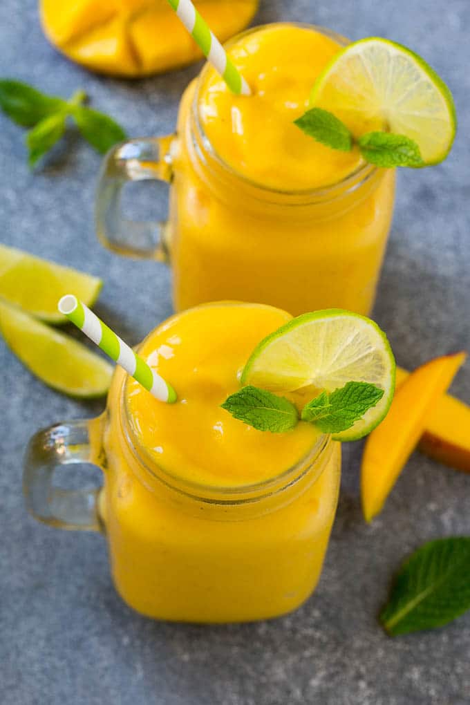 Receta de batido de mango | Receta de mango | Batido saludable #mango #smoothie #drink #dinneratthezoo Dos frascos llenos de batido de mango, aderezado con rodajas de limón y menta fresca.