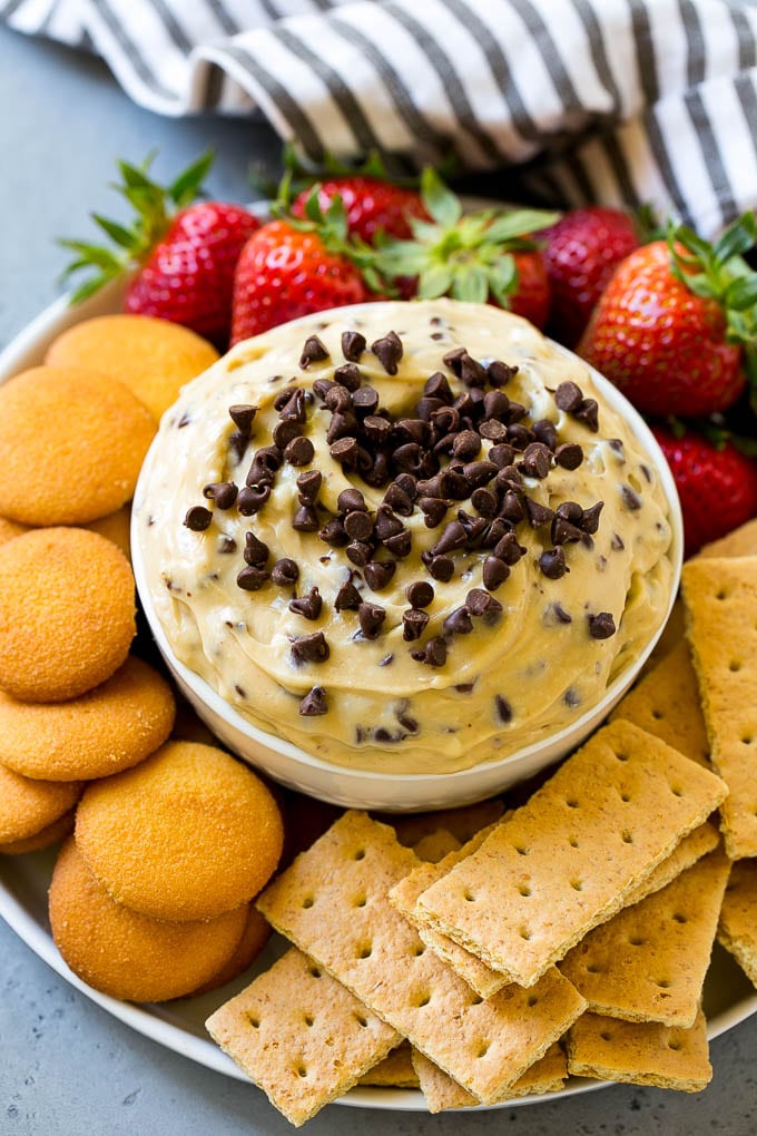 Un tazón de masa para galletas cubierto con chispas de chocolate y servido con galletas y fruta para mojar.