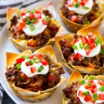 Un plato de tacos hechos con wonton, carne molida sazonada y queso, luego adornado con crema agria, tomates y cebollas verdes.