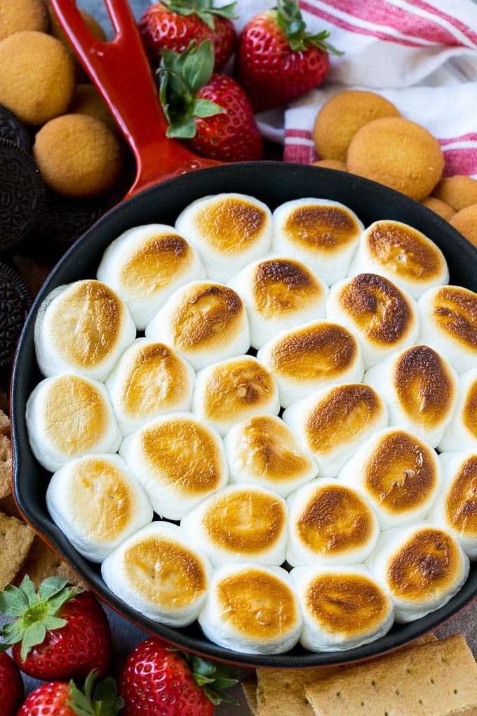 Una sartén de s'mores con chispas de chocolate derretido y malvaviscos tostados, rodeada de galletas saladas, galletas y fresas.