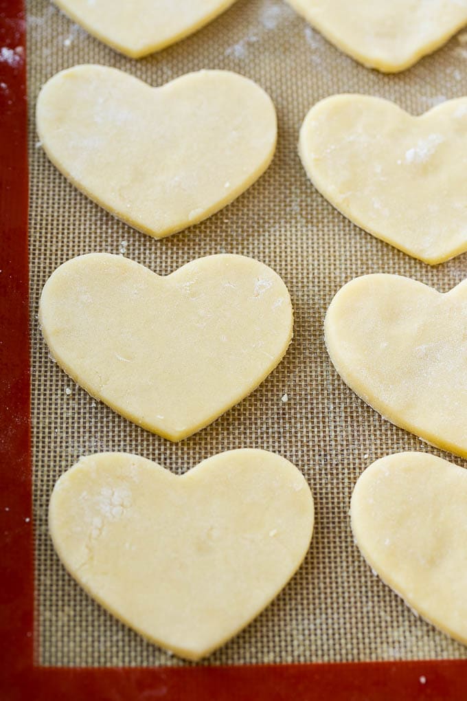 Receta de galletas de corazón | Galletas de azúcar de corazón | Galletas de San Valentín | Galletas de azúcar en forma de corazón | Postre de San Valentín #corazones #cookies #sugarcookies #valentinesday #sprinkles #dessert #dinneratthezoo Trozos de masa de galleta de azúcar en forma de corazón sobre una estera para hornear.