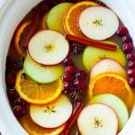 Sidra de manzana en olla de cocción lenta cubierta con rodajas de manzana, naranjas, arándanos, ramas de canela y especias.