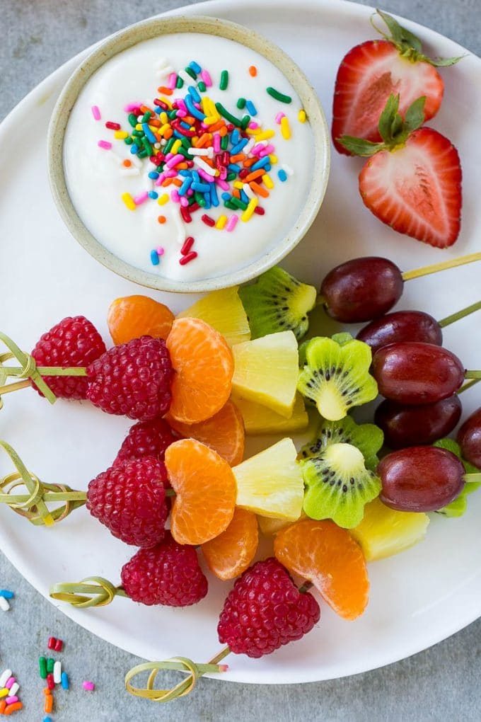 Brochetas de frutas arcoíris en un plato para servir con salsa de yogur.