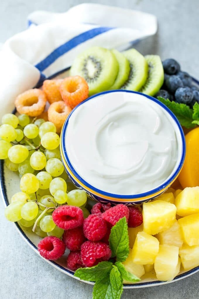 Un tazón de salsa de frutas con queso crema rodeado de frutas coloridas.