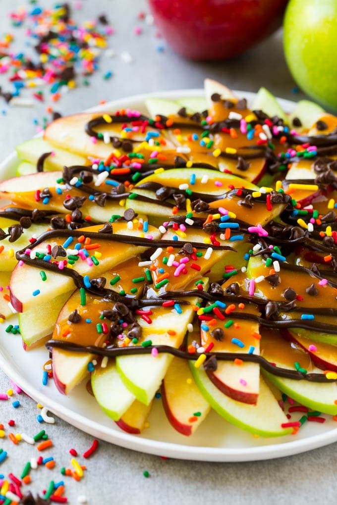 Un plato de manzanas rojas y verdes en rodajas cubiertas con caramelo y chocolate, luego adornado con arco iris y chispas de chocolate.