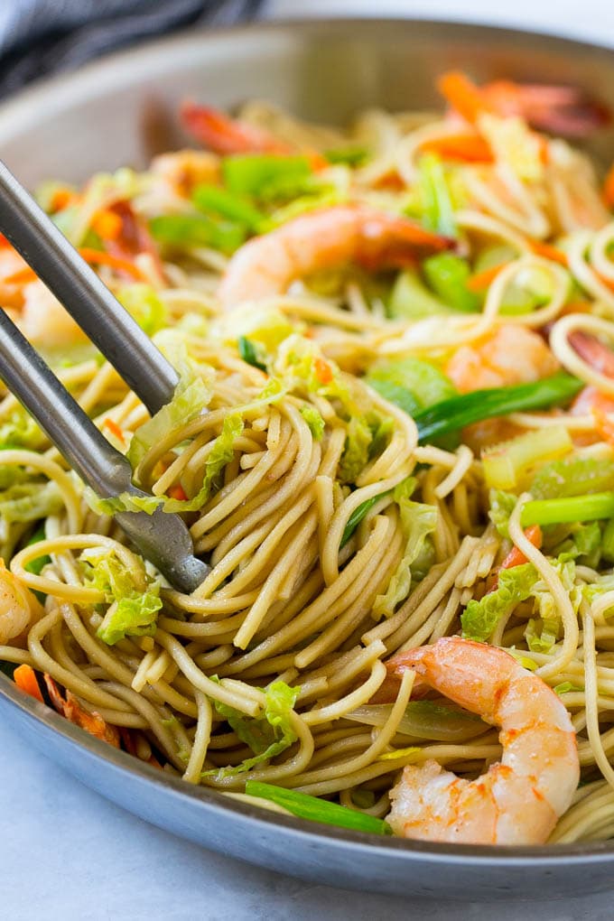 Una sartén llena de camarones chow mein con un par de pinzas levantando los fideos.