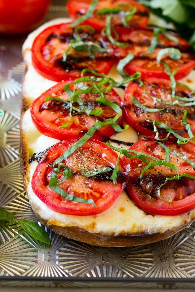 Receta de pan Caprese | Pan de tomate | Pan de queso #caprese #capresebread #paintomate #cheesebread #aperitivo #dinneratthezoo Una hogaza de pan caprese en una bandeja para hornear cubierta con mozzarella fresca, tomates rojos en rodajas, albahaca en rodajas finas y glaseado de vinagre balsámico.