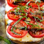 Una hogaza de pan en rodajas en una bandeja para hornear cubierta con mozzarella fresca, tomates rojos en rodajas, albahaca en rodajas finas y glaseado de vinagre balsámico.